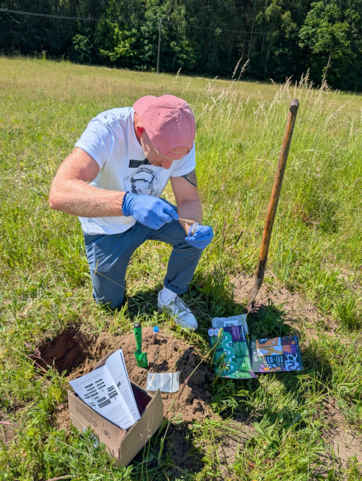 emissionx4 | Charitable Man kneeling in a sunny field, wearing gloves and a pink cap, collecting a soil sample with a small tool beside a shovel and a box of notes.