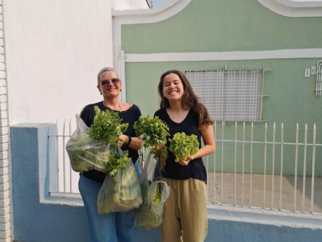 fica4 Two women smiling and holding fresh greens in clear bags outside a pastel-green building with a white fence.
