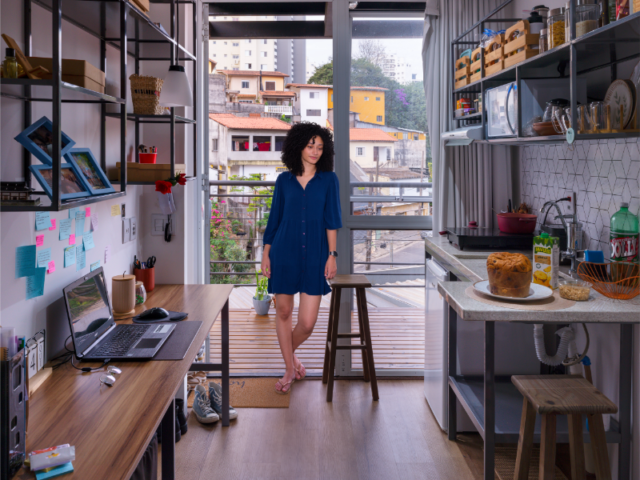 fica5 Woman in a blue dress stands near a balcony door in a compact, modern kitchen area with open shelves and a desk to the left.