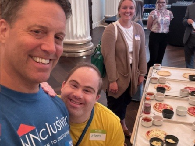 A man in a blue shirt and a shorter man in a yellow shirt smile for a selfie at a buffet table with pizzas and toppings in a formal hall with large white columns in the background.