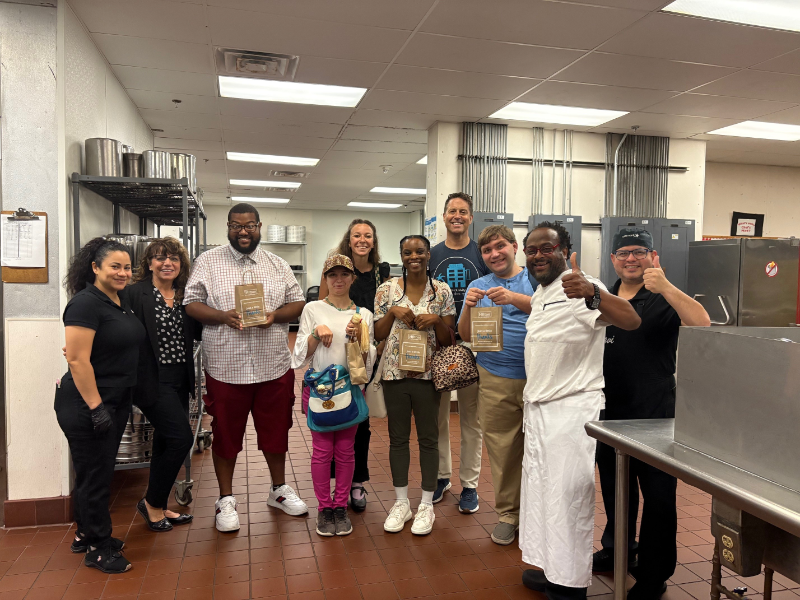 Diverse team of ten in a commercial kitchen posing for a group photo, some holding award plaques and smiling.