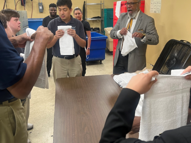 A group of students in a workshop receive towels from a man in a gray suit at a long table in a classroom setting.