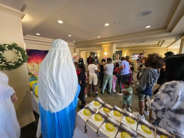 kidsmotelministry3 Crowd gathers in a hall for a celebration; a person in a blue-and-white robe stands near tables of boxed items and a wreath on the wall.
